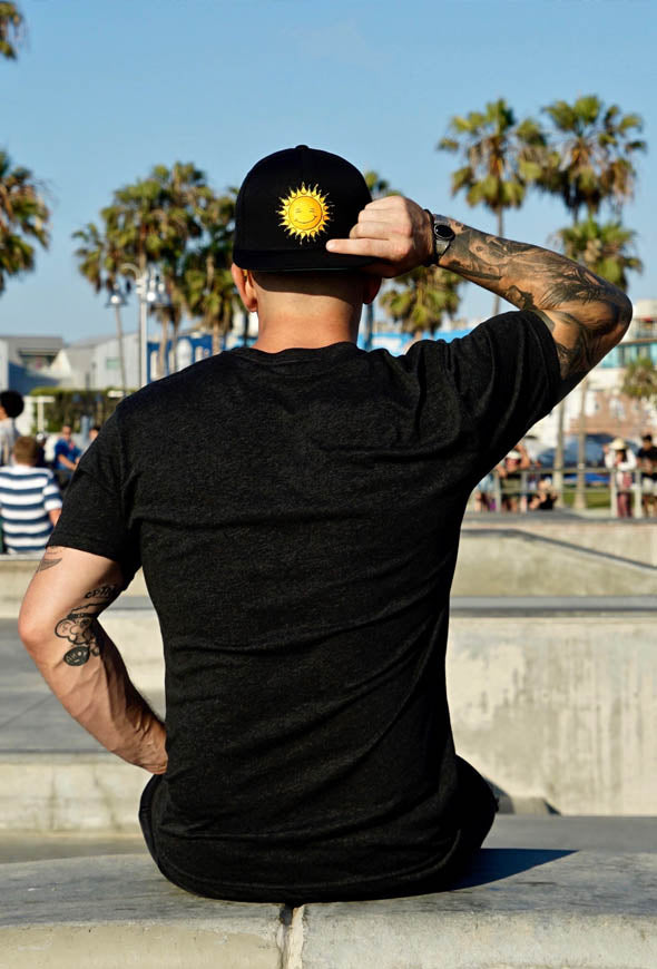 Man facing backwards sitting down on a concrete wall with his new black Kine Threads OG Twill Snapback Hat turned backwards